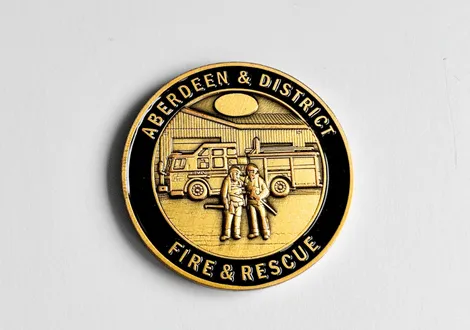 Round badge with black and gold colors featuring two firefighters standing in front of a fire truck and the text 'Aberdeen & District Fire & Rescue'.