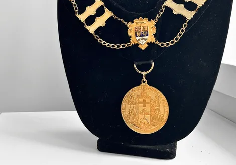 Gold ceremonial medallion with shield emblem and chain displayed on black velvet stand.