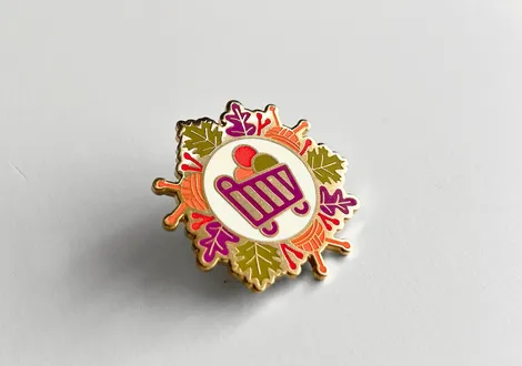 Decorative enamel pin with a central purple shopping cart icon surrounded by autumn leaves in orange, green, and purple colors.