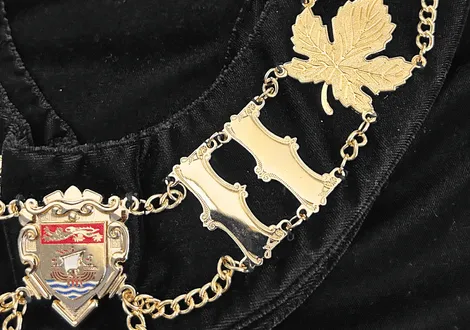 Close-up of a gold chain necklace with a maple leaf and a shield-shaped emblem featuring a sailing ship and red lion design on a black fabric background.