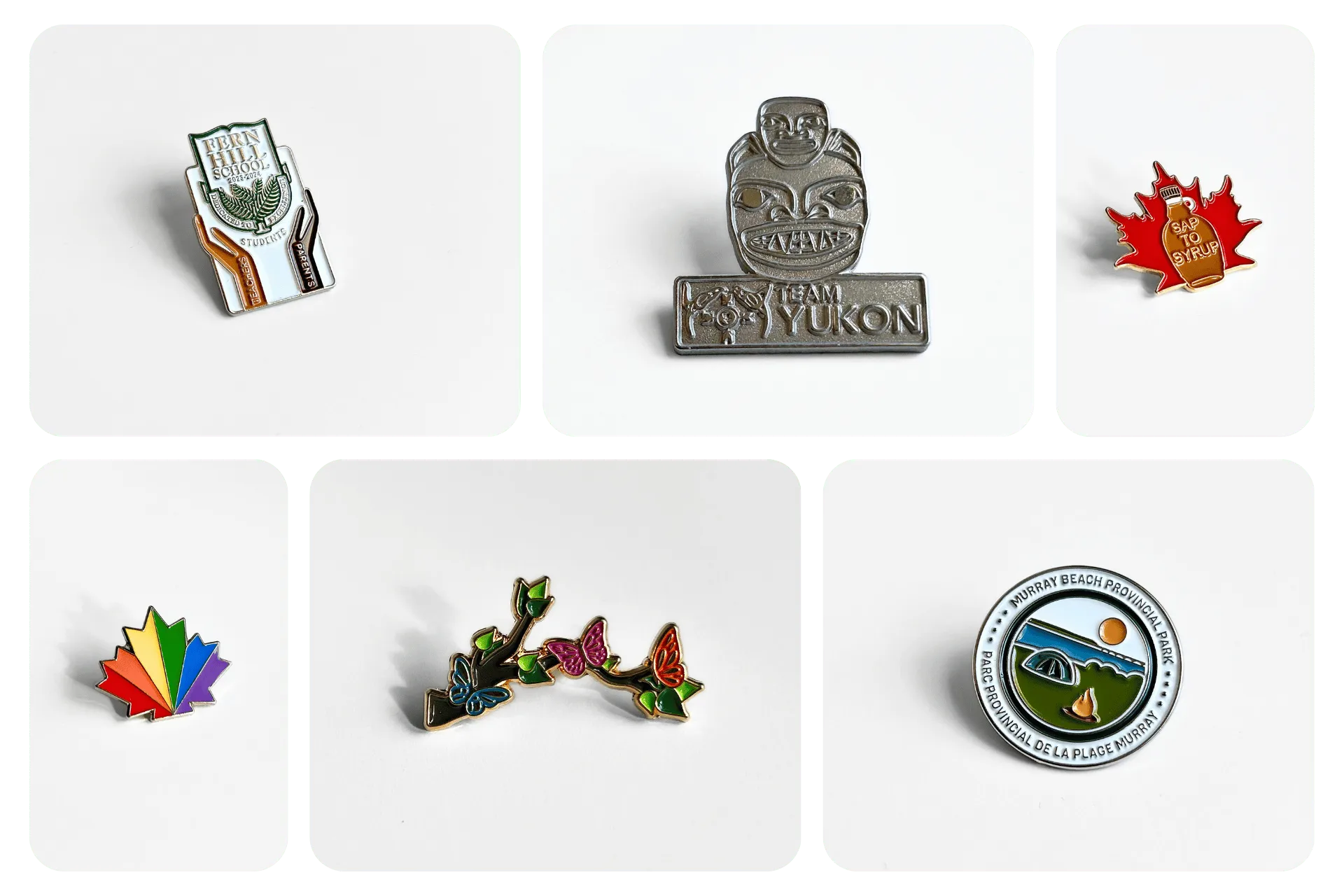 Six enamel pins including a Fern Hill School pin with hands labeled teachers and parents, a Team Yukon silver pin with a traditional mask design, a red maple leaf with a syrup bottle labeled Sap to Syrup, a rainbow maple leaf, a pin of branch with colorful butterflies, and a round Murray Beach Provincial Park pin showing a beach landscape and campfire.