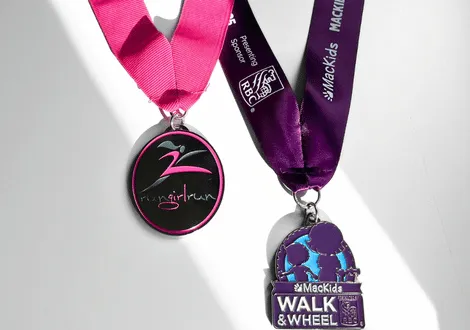Two event medals, one with a pink ribbon and Run Girl Run logo, and the other with a purple ribbon and Mackids Walk & Wheel design.