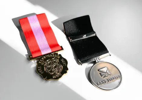 Two medals on a white surface: one with a red and white ribbon and crest, the other a silver round medal with a black ribbon and the text 'LAST TANKS.'