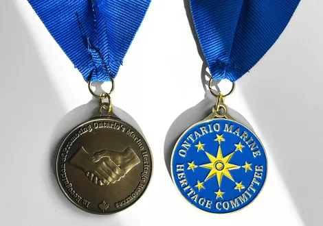Two-sided medal with blue ribbon; one side shows two hands shaking with text honoring Ontario's Marine Heritage Committee, other side features a blue and yellow compass rose with stars and committee name.