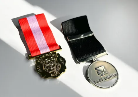 Two medals placed on a white surface, one with a red and white ribbon and the other with a black ribbon, the second medal has 'LAST DANCES' engraved. These are medal examples.