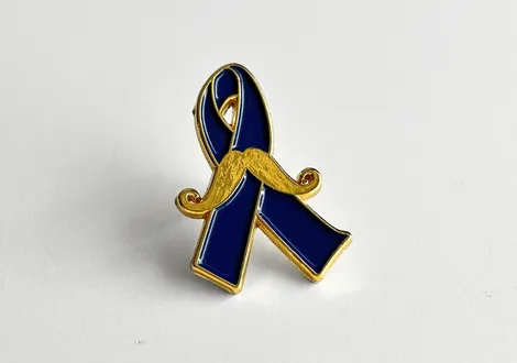 Blue ribbon pin with a gold mustache across the front on a white background.